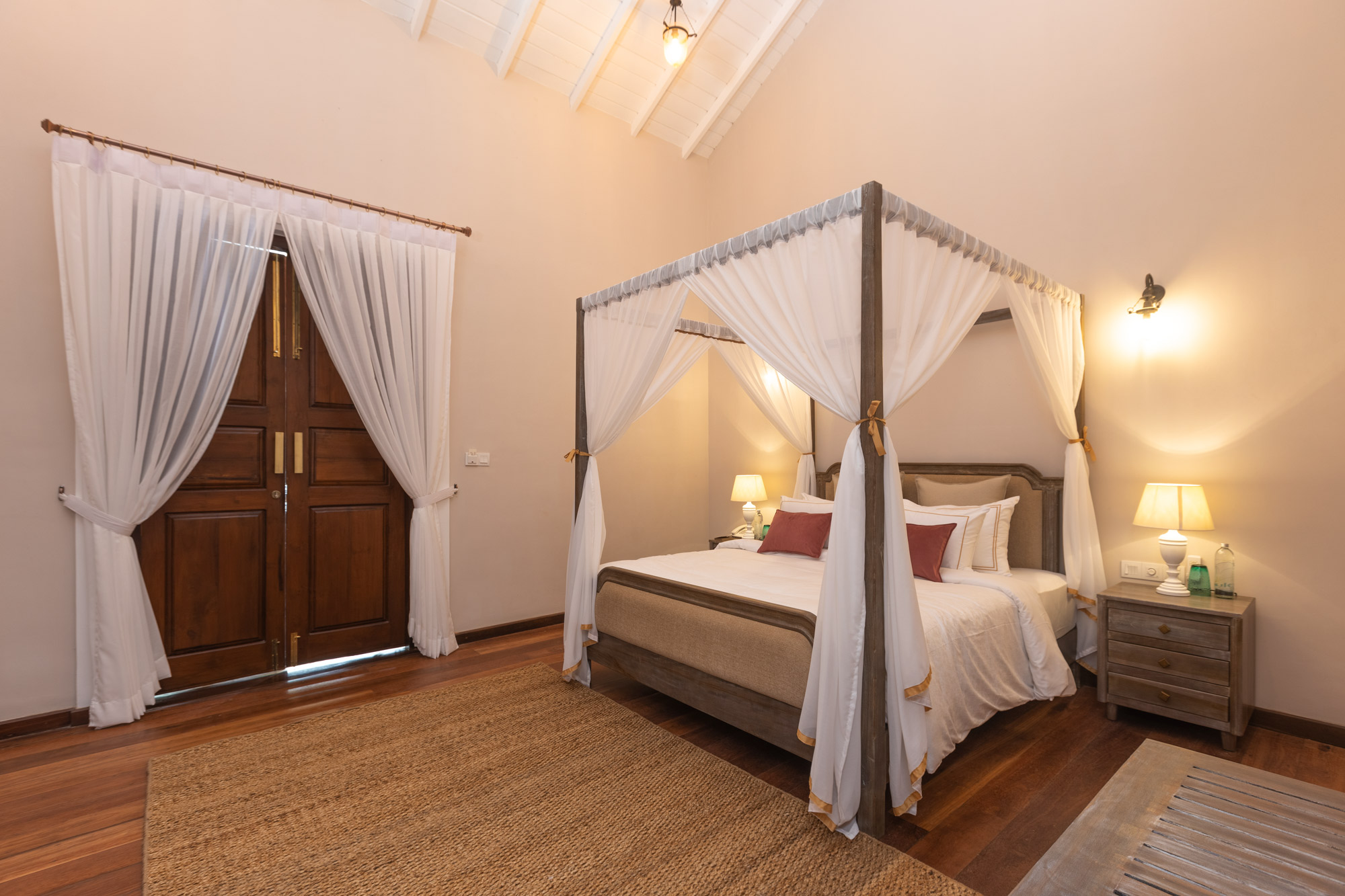A bedroom under a high white-beamed ceiling, with a canopy bed hung with white net curtains, a jute rug on a wooden floor and tall panelled double doors