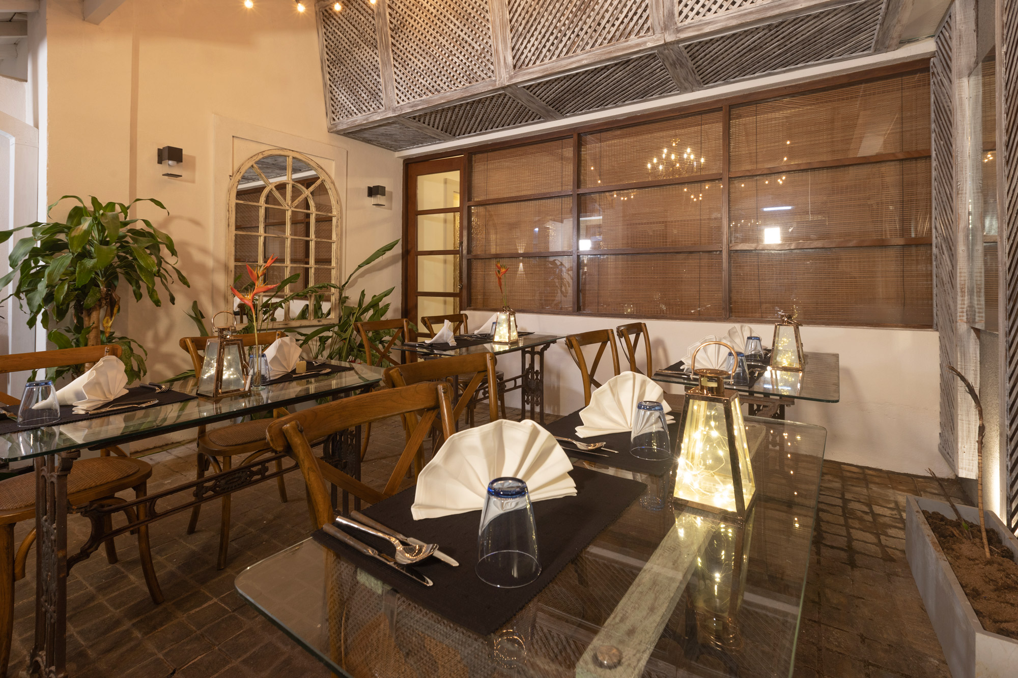 The dining room laid for dinner, with glass-topped tables on iron legs, wooden chairs, folded napkins and lanterns under a timber lattice ceiling