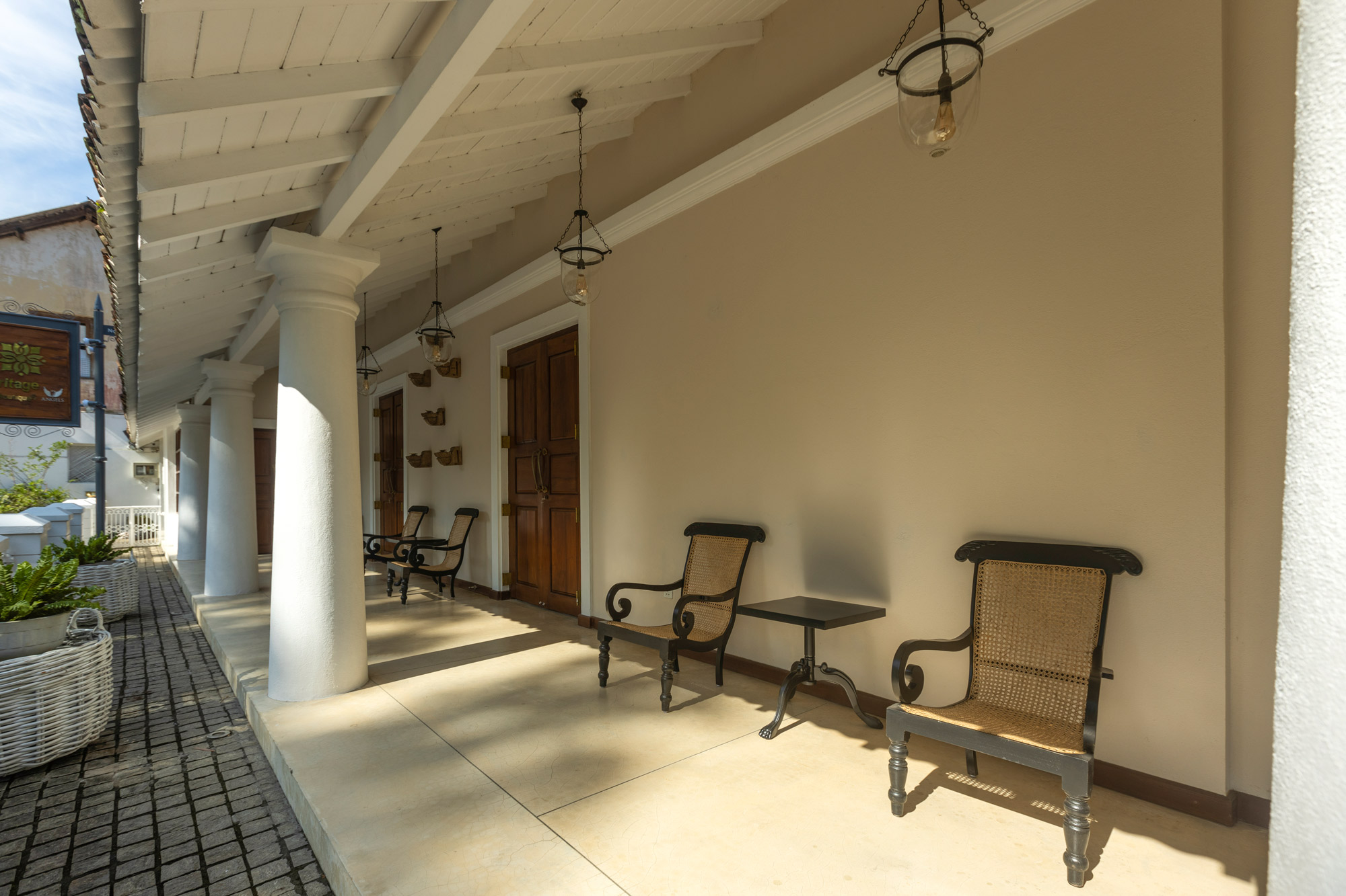 The front veranda in daylight: white columns, hanging glass lanterns, wooden doors and cane planters' chairs along the wall
