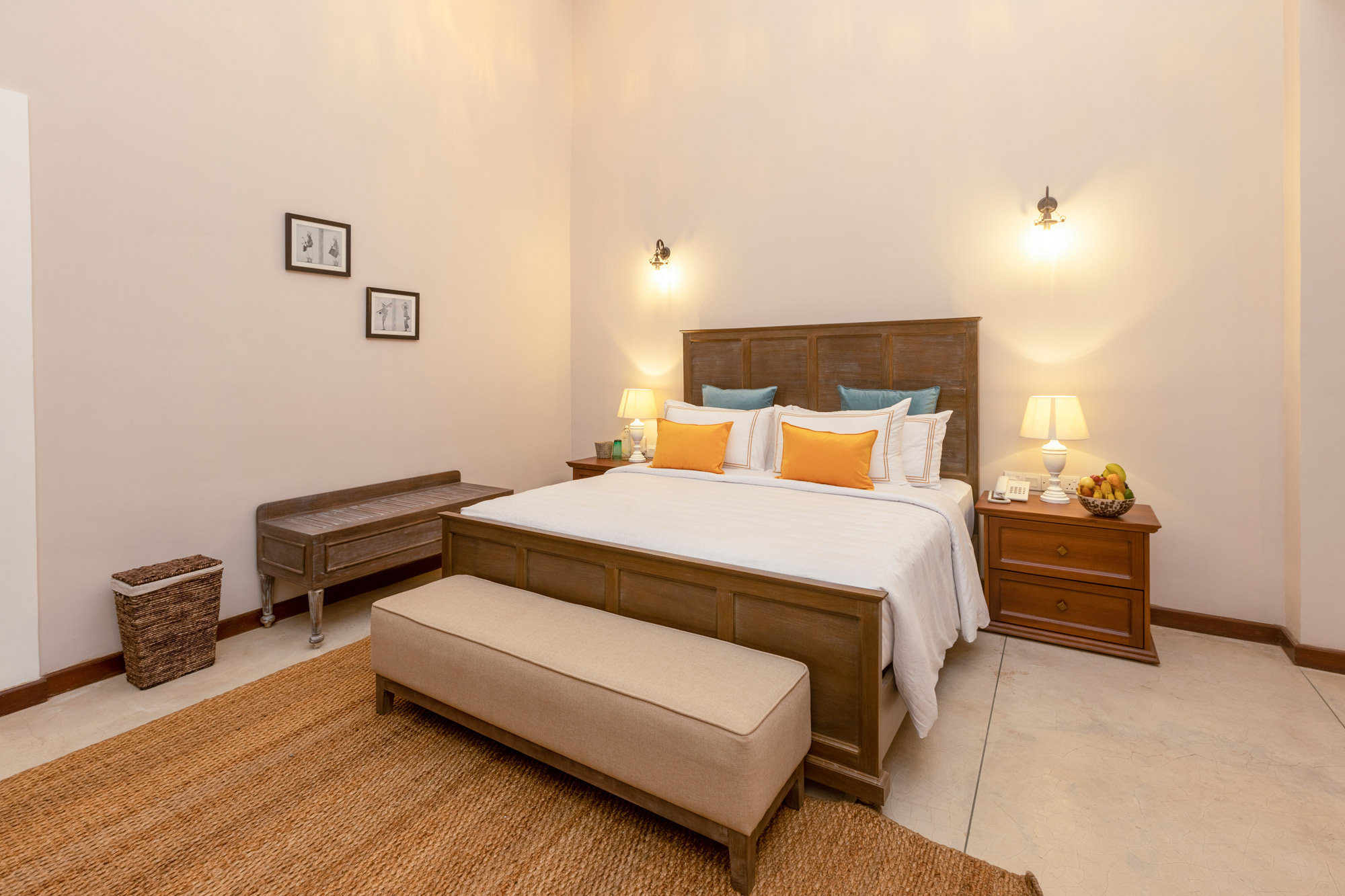 A bedroom with a panelled wooden headboard, a double bed with orange and blue cushions, an upholstered bench at its foot and a jute rug